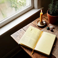 Open journal on a wooden surface with a lit candle and small plants near a window.